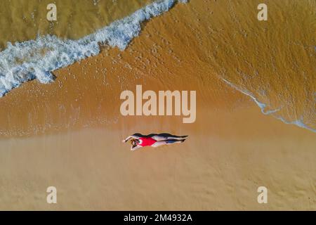 Femme en maillot de bain rouge couché sur la belle plage de sable avec vagues de l'océan, vue aérienne de drone. Banque D'Images