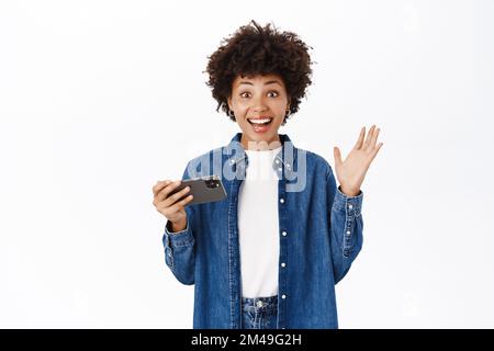 Une jeune femme heureuse et belle saute de l'excitation, en utilisant un téléphone mobile et en regardant joyeusement, en gagnant sur l'application pour smartphone, debout sur fond blanc Banque D'Images