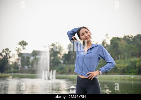 Femme asiatique millénaire attrayante et sportive en vêtements de sport qui s'étire le cou et s'échauffe avant de courir dans le magnifique parc. style de vie et activité Banque D'Images