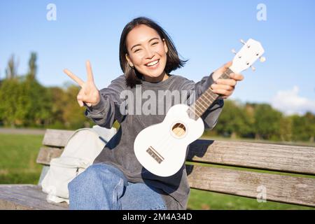 Happy asian girl montre ukulele et pouces vers le haut, montre son nouvel instrument de musique, apprend à jouer dans le parc, assis sur le banc Banque D'Images