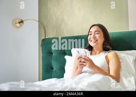 Belle femme asiatique souriante au lit, en lisant des messages sur un smartphone et en riant, en profitant d'une journée de repos dans la chambre, à l'aide d'un téléphone portable Banque D'Images