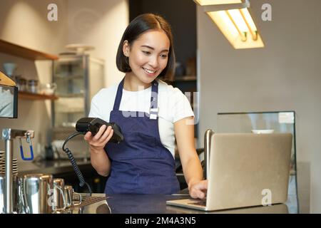 Un barista asiatique souriant, une fille avec un terminal de carte, une machine de paiement et un ordinateur portable, debout dans un café, traitant le paiement de la commande de café Banque D'Images