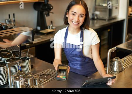 Portrait de la belle fille asiatique Barista, le personnel du café-restaurant donne le terminal de PDV, aide à payer sans contact avec la carte de crédit, debout au comptoir dans le café Banque D'Images