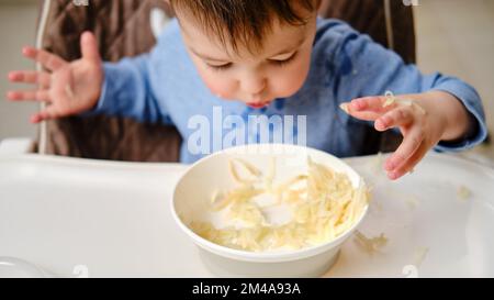 Drôle d'enfant manger une pomme râpée avec sa main d'une assiette, gros plan. Bébé garçon affamé mange des fruits, de l'humour. Enfant âgé d'un an et trois mois Banque D'Images