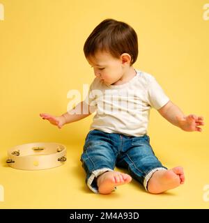 Bébé tout-petit joue le tambourin, un enfant avec un instrument de musique à percussion sur fond jaune studio. Joyeux musicien d'enfant jouant le tambour de main. Banque D'Images
