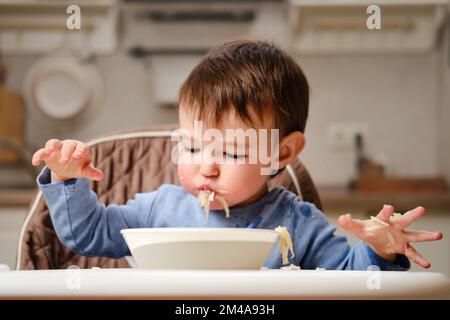 Un enfant drôle mange une pomme râpée avec sa bouche pleine tout en étant assis sur une chaise de cuisine. Bébé garçon affamé, il a de la nourriture dans sa bouche, de l'humour. Enfant âgé Banque D'Images