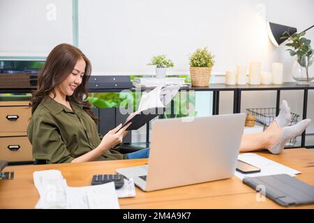 Femme d'affaires assise relaxante inclinable avec les pieds sur le bureau d'ordinateur lire le rapport Banque D'Images