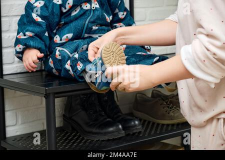 La mère met des chaussures bleues sur le pied de bébé enfant assis dans le couloir de la maison. Femme maman vêtue chaud bottes vêtements sur enfant pour l'hiver marcher dans c Banque D'Images