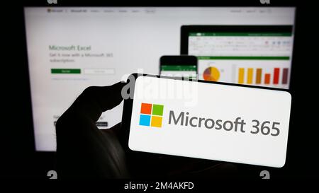 Personne tenant un téléphone portable avec le logo du logiciel de productivité Microsoft 365 à l'écran en face de la page Web de l'entreprise. Mise au point sur l'affichage du téléphone. Banque D'Images