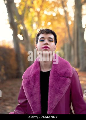 Auto-assuré jeune homme liquide de genre avec cheveux courts et maquillage dans le cuir fuchsia brillant et manteau de fourrure debout près des arbres d'automne et regardant le chamer Banque D'Images