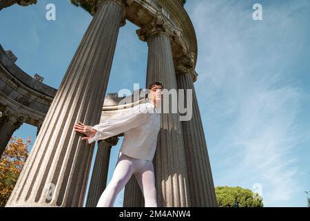 De dessous de l'extravagant homme avec les cheveux courts et le maquillage dans la chemise blanche et les bas debout près des colonnes hautes contre ciel bleu ciel nuageux le jour ensoleillé Banque D'Images