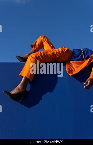 Vue latérale en contre-plongée d'une femme afro-américaine non reconnaissable, en costume orange et talons hauts posés sur un mur bleu et bénéficiant d'un temps ensoleillé Banque D'Images