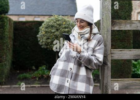 Bonne jeune femme en vêtement d'extérieur élégant regardant la vidéo sur smartphone tout en se penchant sur la clôture de bois contre la vieille maison et la haie luxuriante à Édimbourg, Écosse Banque D'Images