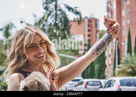 Confiante jeune femme optimiste avec de longs cheveux ondulés blond et tatouage sur les bras dans des lunettes de soleil stylées agitant la main et souriant heureux tout en prenant selfi Banque D'Images