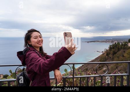 Jeune femme souriante vêtue de vêtements chauds debout sur la terrasse et se faisant autoportrait contre la vue pittoresque de la mer pendant le voyage à Taormina Banque D'Images