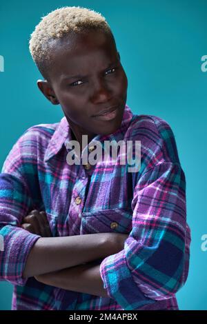 Une jeune Afro-américaine mécontente, en chemise à carreaux, avec des cheveux bouclés blonds croisant les bras et regardant la caméra sur fond bleu Banque D'Images