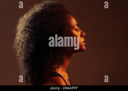 Vue latérale d'une femme afro-américaine pensive vêtu d'une robe colorée qui se tient debout dans un studio sombre, près d'un fond marron Banque D'Images