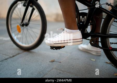Anonyme Afro-américaine femme en baskets blanches marcher sur la pédale de vélo tout en se tenant sur la chaussée sur la rue de la ville Banque D'Images