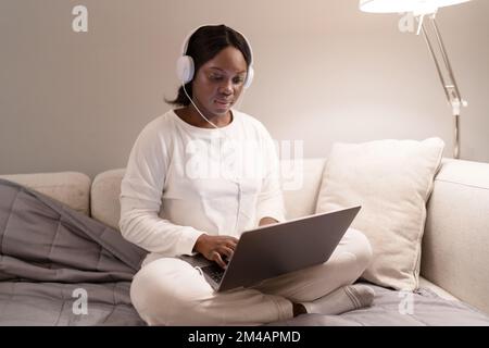 Jeune Afro-américaine en pyjama blanc et chaussettes avec casque pour regarder un film sur un ordinateur portable tout en étant assise à pieds croisés sur un canapé dans le salon à Banque D'Images