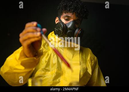 Homme en costume noisette et masque à gaz utilisant une pipette pour ajouter le réactif rouge dans le flacon tout en faisant des drogues illégales dans le laboratoire sombre Banque D'Images