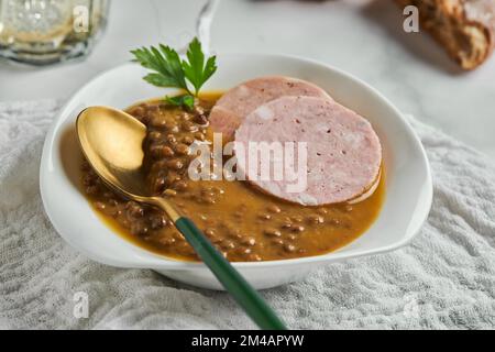 De dessus cuillère placée dans un bol de soupe de lentilles délicieuses servi avec des tranches de saucisse et une feuille de persil sur table en marbre et serviette grise Banque D'Images