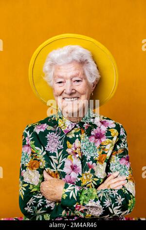Une femme d'âge heureuse en chemise fleurie et chapeau feutre croisant les bras et regardant l'appareil photo avec le sourire sur fond jaune Banque D'Images
