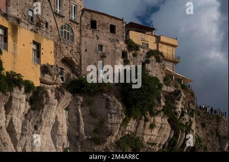 Fondations rocheuses : de vieilles maisons en ruines de pierre partiellement rendues semblent s'accrocher au bord abrupt des falaises verticales de grès qui soutiennent la vieille ville de Tropea, sur la côte ouest de la Calabre, dans le sud de l'Italie, tandis que les visiteurs admirent le paysage spectaculaire en contrebas d'un belvédère ou d'un point de vue voisin. Banque D'Images