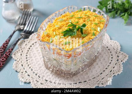 Salade de fête Mimosa avec poisson en conserve, pommes de terre, fromage, carottes et œufs dans un saladier transparent sur fond bleu Banque D'Images