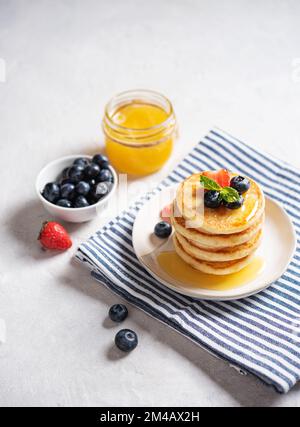 Crêpes américaines classiques faites maison dans une pile de baies fraîches et de miel sur une serviette rayée sur un fond de texture gris clair. Le concept d'un He Banque D'Images