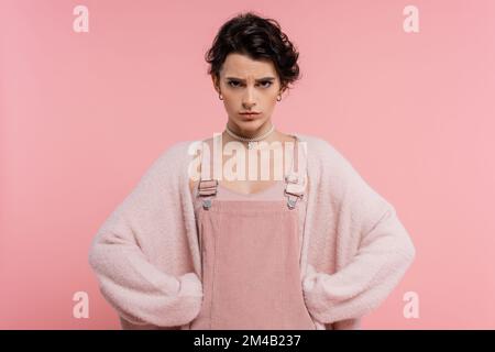 femme en colère dans la robe de sangle et gilet chaud debout akimbo et regardant l'appareil photo isolé sur rose Banque D'Images