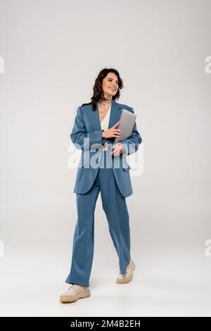 pleine longueur de femme souriante en costume bleu marchant avec ordinateur portable sur fond gris Banque D'Images