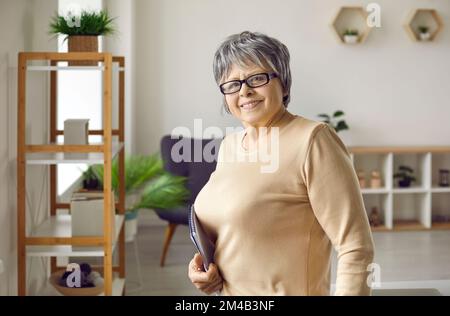 Portrait d'une femme âgée souriante avec bloc-notes à la main dans le salon de sa maison. Banque D'Images