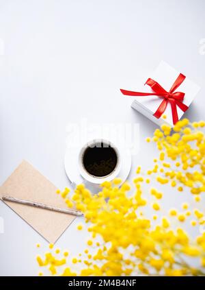 un bouquet de printemps avec des fleurs de mimosa jaunes sur une tasse de café noir, une boîte de cadeau et un cahier artisanal sur une table blanche. Concept du 8 mars Banque D'Images