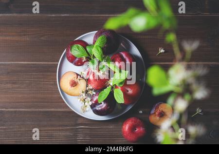 quelques prunes rouges juteuses dans une assiette bleue avec des fleurs de branches sur une serviette sur un fond de bois brun. Vue de dessus et image d'espace de copie Banque D'Images