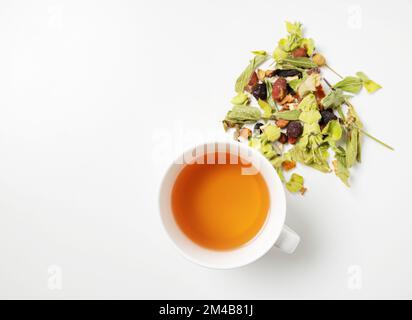 Une tasse de tisane avec des herbes séchées éparpillées et des morceaux de fruits sur un fond blanc. Vue de dessus et espace de copie. Banque D'Images