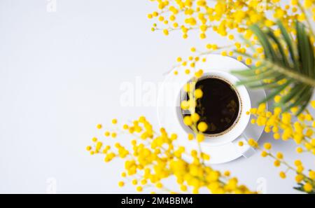 un bouquet de printemps avec des fleurs de mimosa jaunes sur une tasse de café noir sur une table blanche. Concept du 8 mars, bonne journée des femmes. Vue de dessus et copie de la flèche Banque D'Images