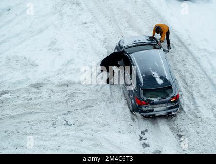 Deux hommes poussent une voiture coincée dans la neige. La voiture glisse dans une dérive de neige sur une route glacée. Routes non nettoyées de la neige. Banque D'Images