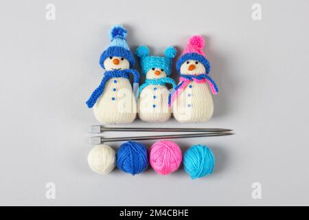 Trois bonhommes de neige tricotés en chapeaux bleu et rose avec foulards avec boules de fil et aiguilles de tricotage sur fond gris. Créativité de tricot d'hiver, t Banque D'Images