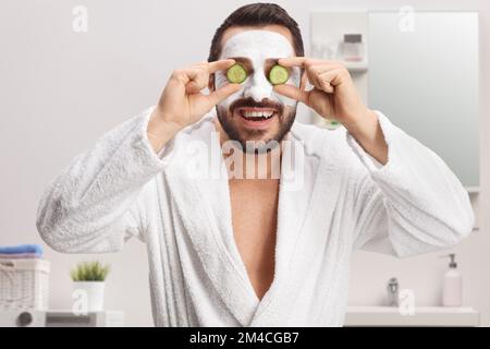 Homme dans un peignoir avec un masque facial tenant des concombres sur les yeux dans une salle de bains à la maison Banque D'Images