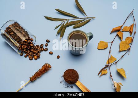 La vie dure avec du café moulu au premier plan, des grains de café derrière et une tasse à l'arrière-plan, des feuilles sèches assorties et une surface bleue lisse Banque D'Images