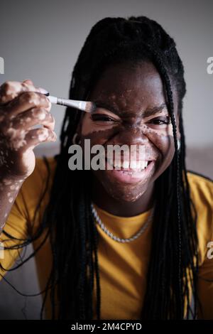 Positive noire femme avec pigmentation blanche riant tout en faisant le maquillage Banque D'Images