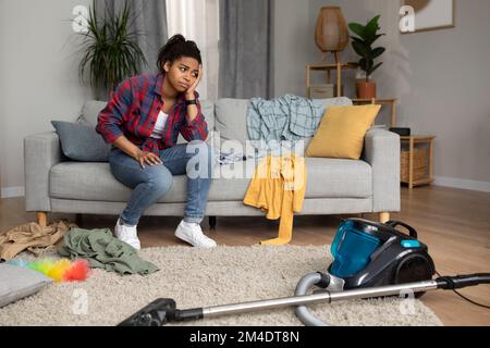 Une femme afro-américaine triste et fatiguée s'assoit sur un canapé, regarde l'aspirateur et souffre du chaos et de la pagaille Banque D'Images