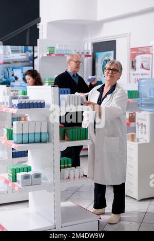 Pharmacien âgé caucasien debout en pharmacie arranger des étagères tout en vérifiant des paquets de pilules, aidant les clients avec des prescriptions de soins de santé. Service et support médicaux Banque D'Images