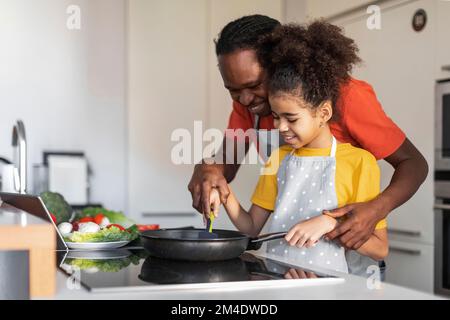 Temps de cuisson. Joyeux papa noir et fille de préadolescence préparant le déjeuner dans la cuisine Banque D'Images