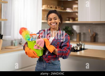 Joyeuse femme afro-américaine millénaire en gants de caoutchouc avec des fournitures de nettoyage seau aime les tâches ménagères Banque D'Images