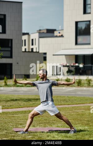 Portrait vertical complet d'un jeune homme faisant du yoga à l'extérieur en plein soleil dans un complexe de logement moderne Banque D'Images