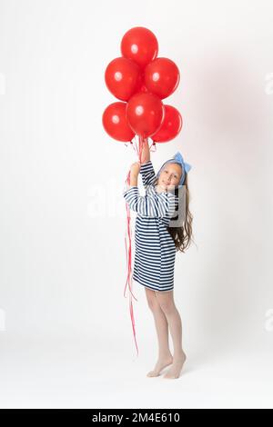 Fille tenant un grand bouquet de ballons rouges dans les mains, souriant et regardant l'appareil photo. Studio photo du modèle 10 ans en robe à rayures blanches et bleues Banque D'Images