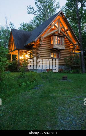 maison en rondins de style chalet construite en 2003 avec un toit de bardeaux d'asphalte brun et une fondation en pierre naturelle illuminée au crépuscule au printemps. Banque D'Images