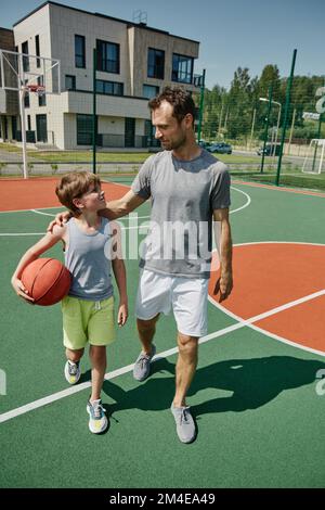 Portrait vertical complet de père et fils heureux jouant au basket-ball ensemble sur un terrain de sport éclairé par la lumière du soleil Banque D'Images