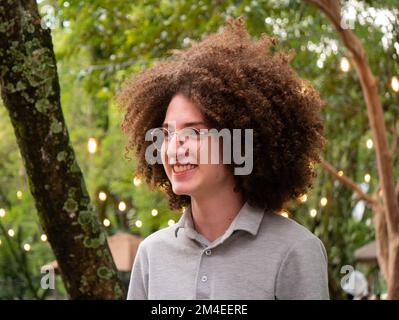 Medellin, Antioquia, Colombie - 14 novembre 2022: Jeune homme mince avec cheveux et lunettes de curly se tient dans un parc plein d'arbres Banque D'Images